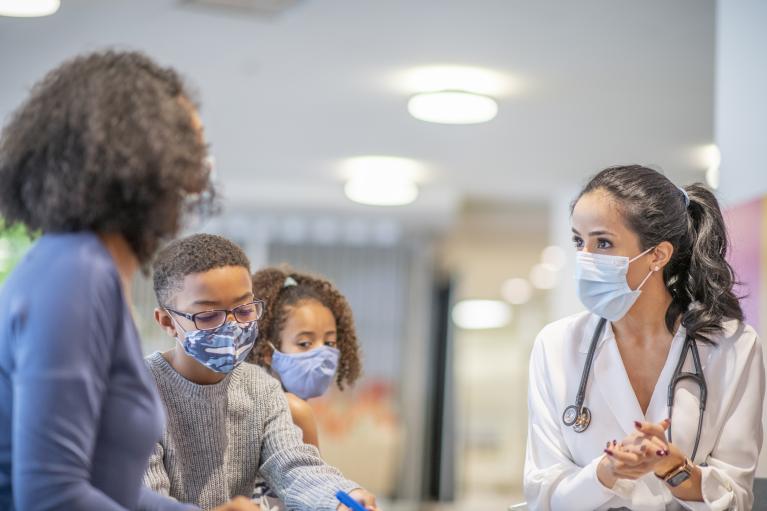 A doctor interacts with a mother and her two children in a facility. They are all wearing masks.